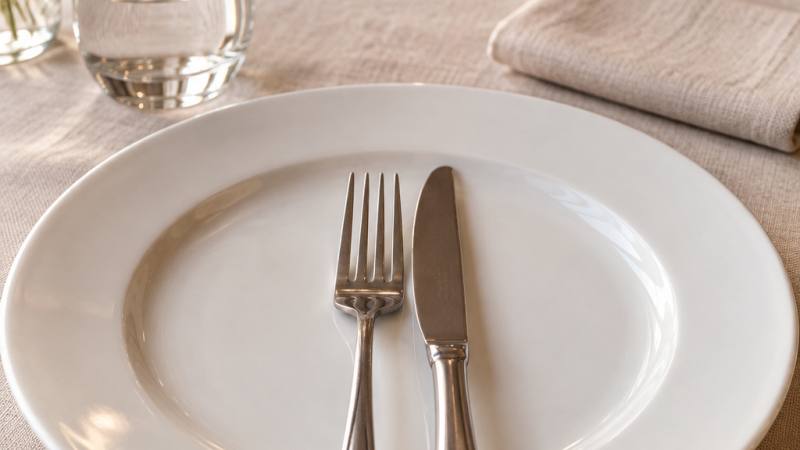 Assiette blanche avec fourchette et couteau posés parallèlement, lame tournée vers l’intérieur, sur une nappe de lin